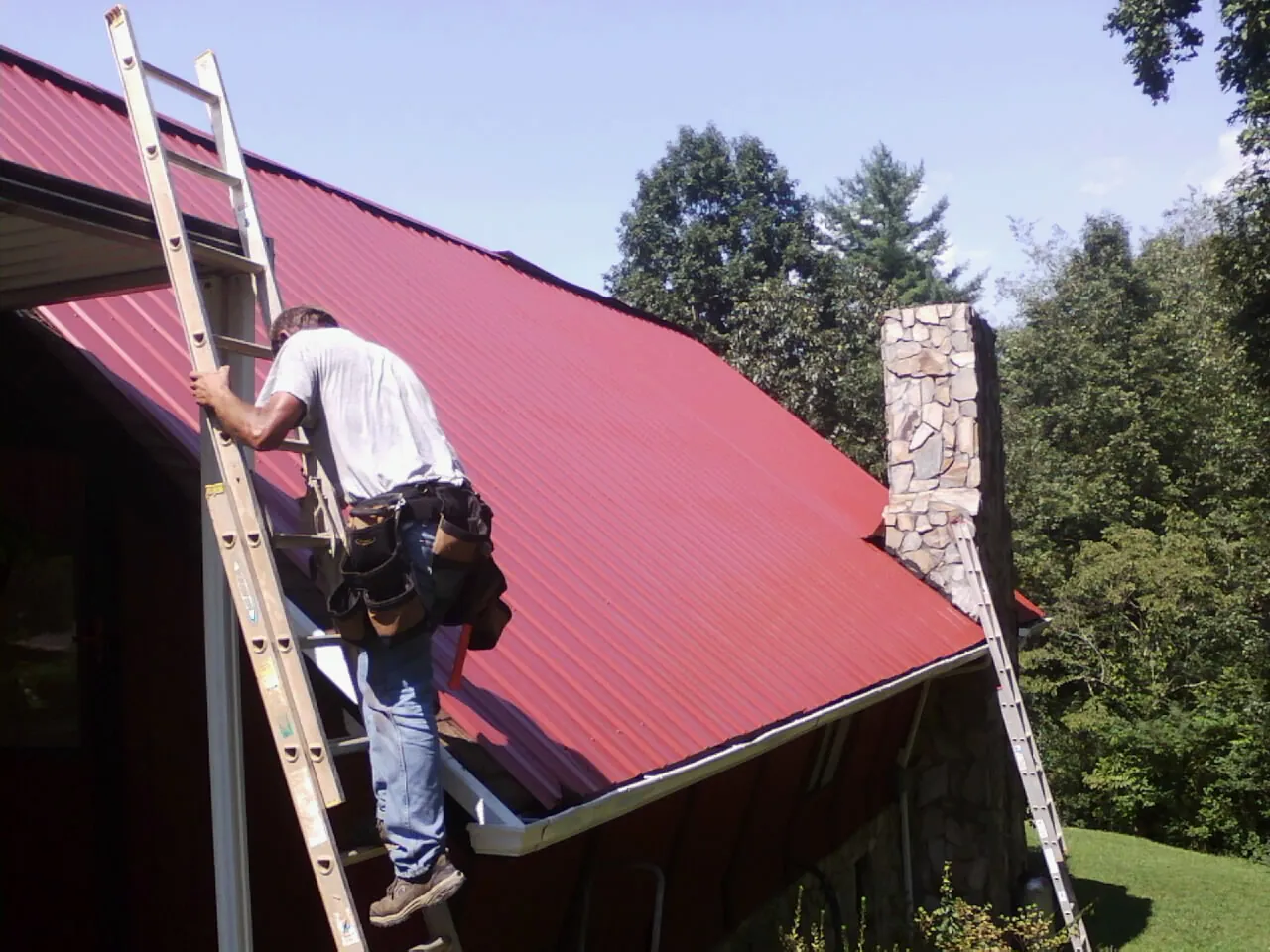 Roofer on a ladder beside a red metal roof and stone chimney — Soffit & Fascia Repair by Roofing Brothers in Richton Park